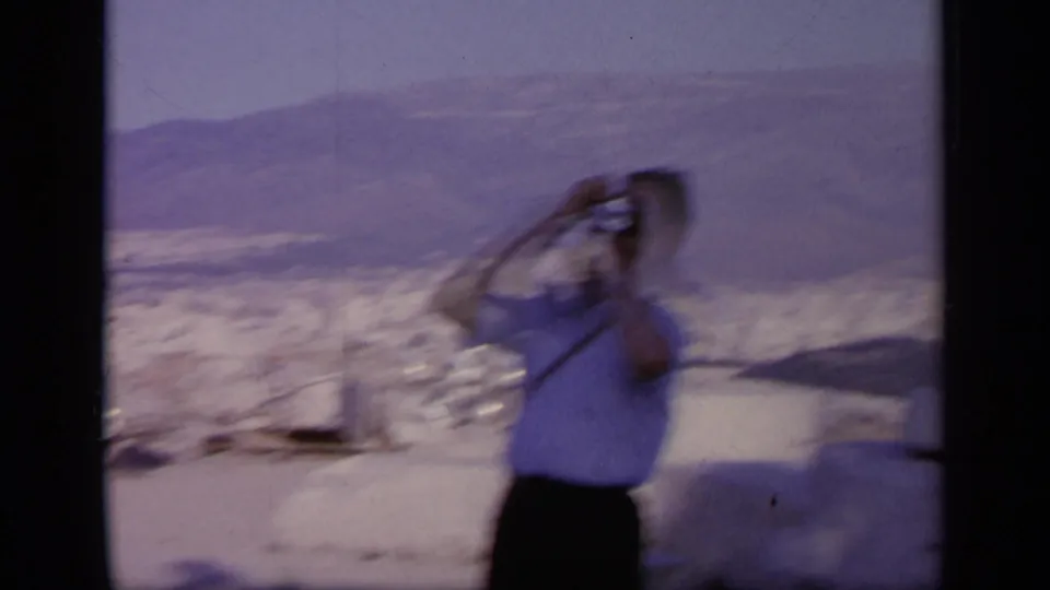 1963 Greece home-movie frame with a blurred tourist lifting a camera, backed by hills and pale stone ruins.
