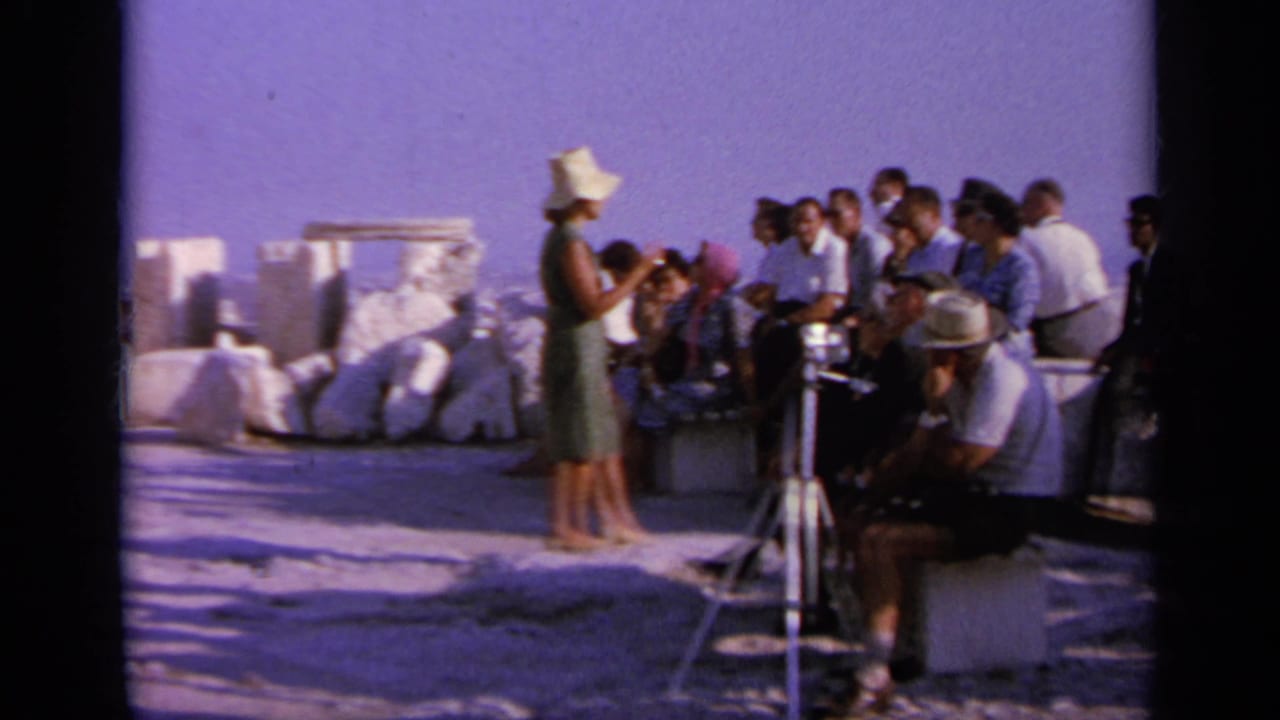1963 Greece frame showing tourists gathered by ancient-looking stone remains while a woman in a hat stands near a tripod camera.