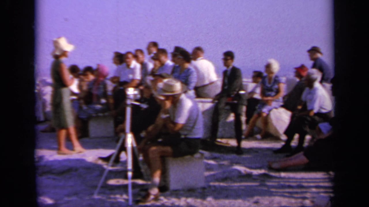 Crowd gathered near a tripod camera at a sunlit stone site.
