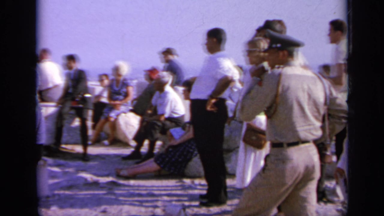 Visitors and uniformed men stand near a seated crowd at a Greek archaeological site in 1963 home-movie footage.