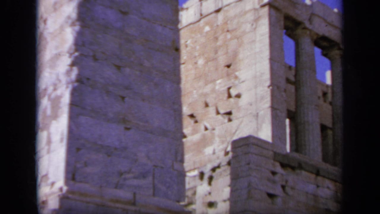 The camera cuts to weathered stone masonry and window-like openings at a Greek ruin site.