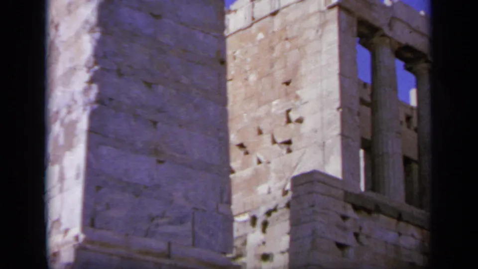 The camera cuts to weathered stone masonry and window-like openings at a Greek ruin site.