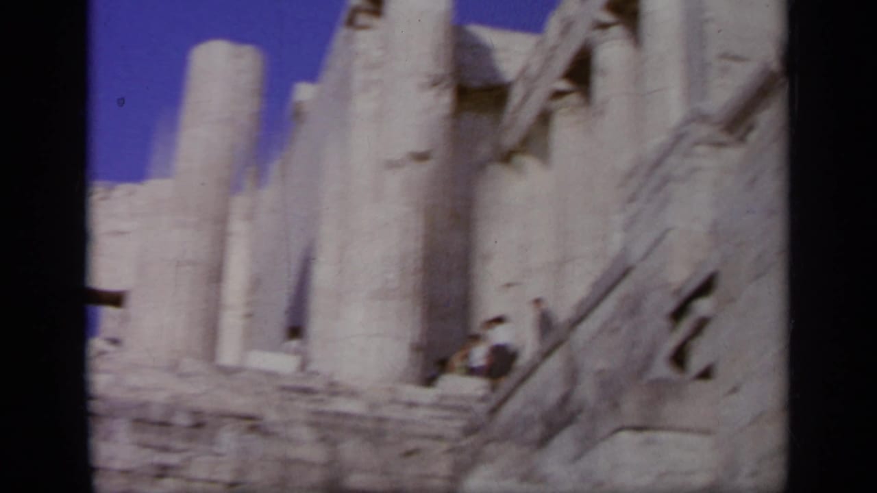 Handheld 1963 Greece frame looking across columns, stone steps, and a distant visitor at an ancient ruin site.