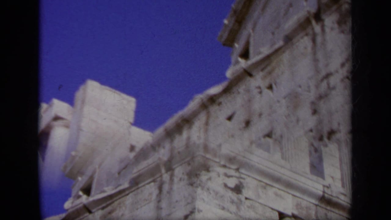 1963 Greece home-movie frame angled upward toward a weathered ruin wall, columns, and open blue sky.