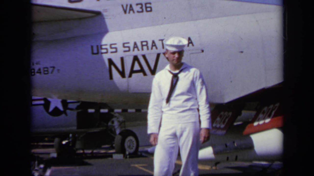 Young sailor in white uniform standing beside a Navy aircraft marked USS Saratoga.