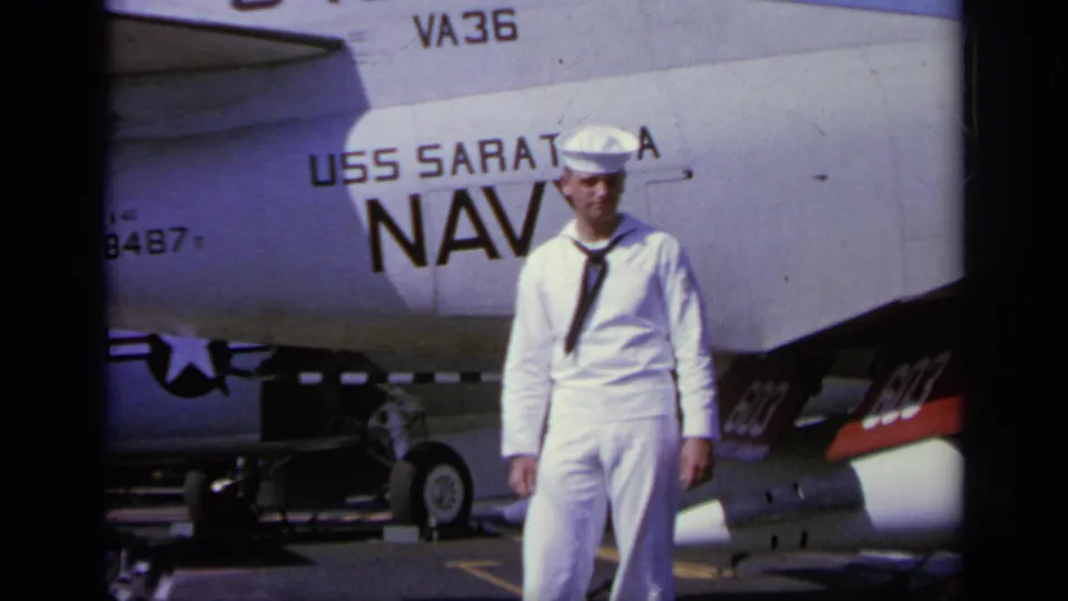 Young U.S. Navy sailor in white uniform posing on a flight deck beside aircraft lettering for USS Saratoga and NAVY.