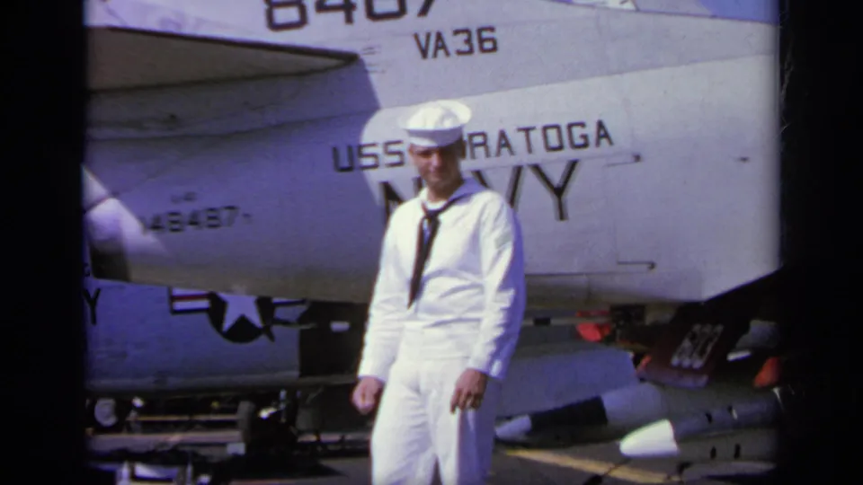 1963 home-movie frame of a U.S. Navy sailor standing close to aircraft markings reading USS Saratoga, NAVY, and VA-36.