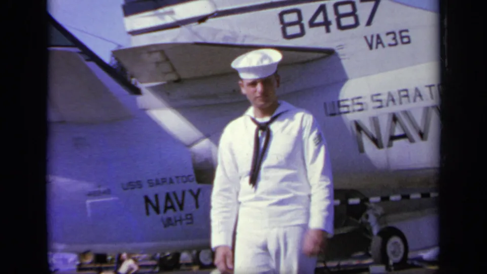 U.S. Navy sailor on a carrier flight deck beneath aircraft number 8487, with USS Saratoga and NAVY markings visible.