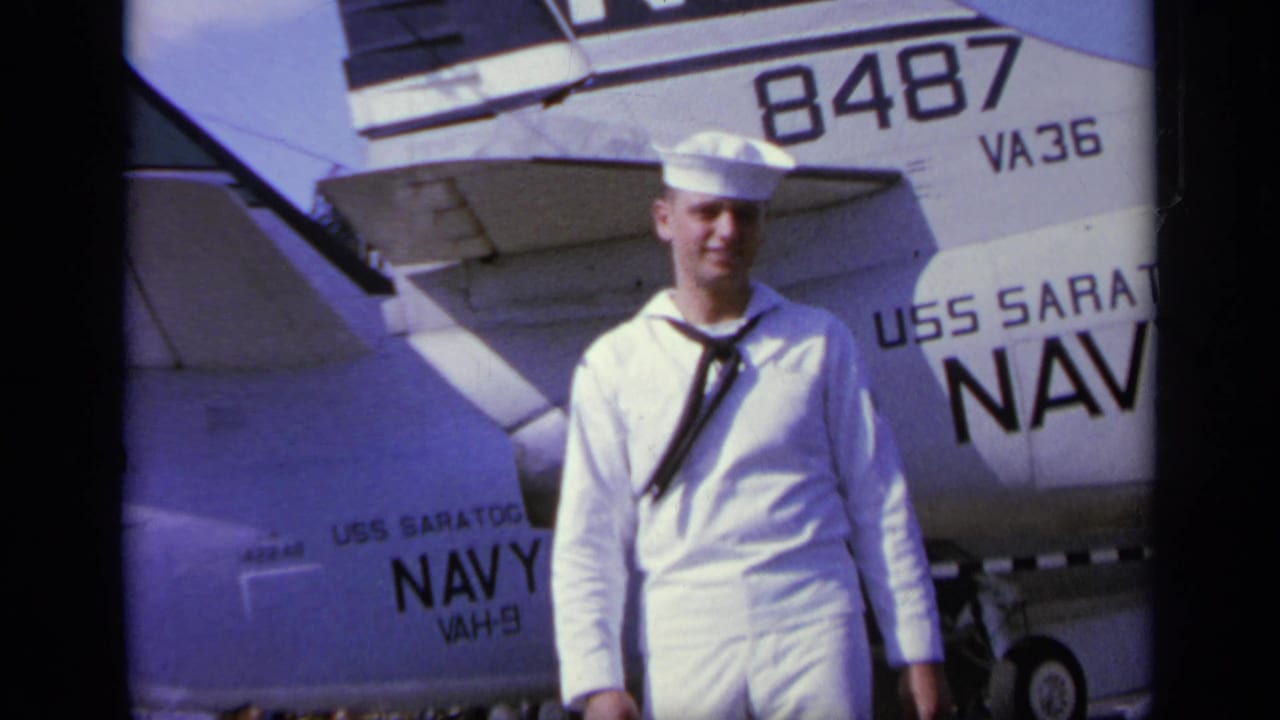 In Greece in 1963, a young sailor in dress whites smiles on the USS Saratoga flight deck beside a Navy aircraft tail marked 8487 and VA36.