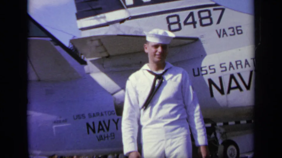 In Greece in 1963, a young sailor in dress whites smiles on the USS Saratoga flight deck beside a Navy aircraft tail marked 8487 and VA36.