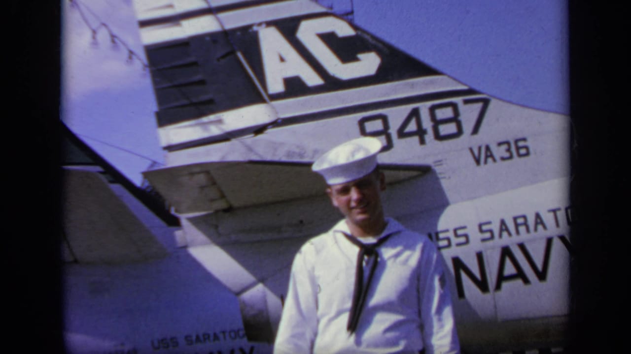A sailor in dress whites stands on the USS Saratoga flight deck near aircraft tail markings including AC, 9487, and VA36.