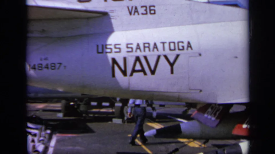 Close 8mm view of USS Saratoga and NAVY lettering on an aircraft fuselage, with deck equipment and a partial crew figure below.