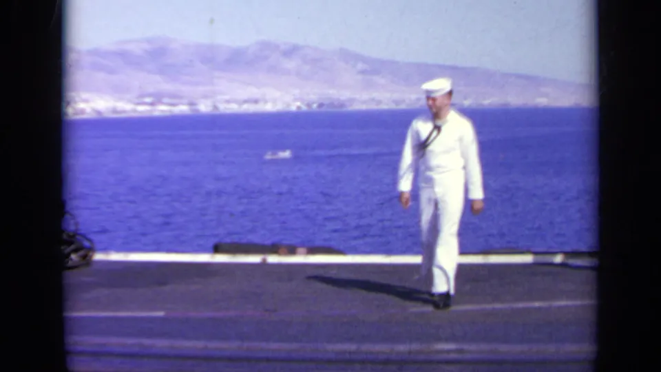 A sailor in dress whites walks across the USS Saratoga flight deck in Greece, with blue water and distant mountains in the background.