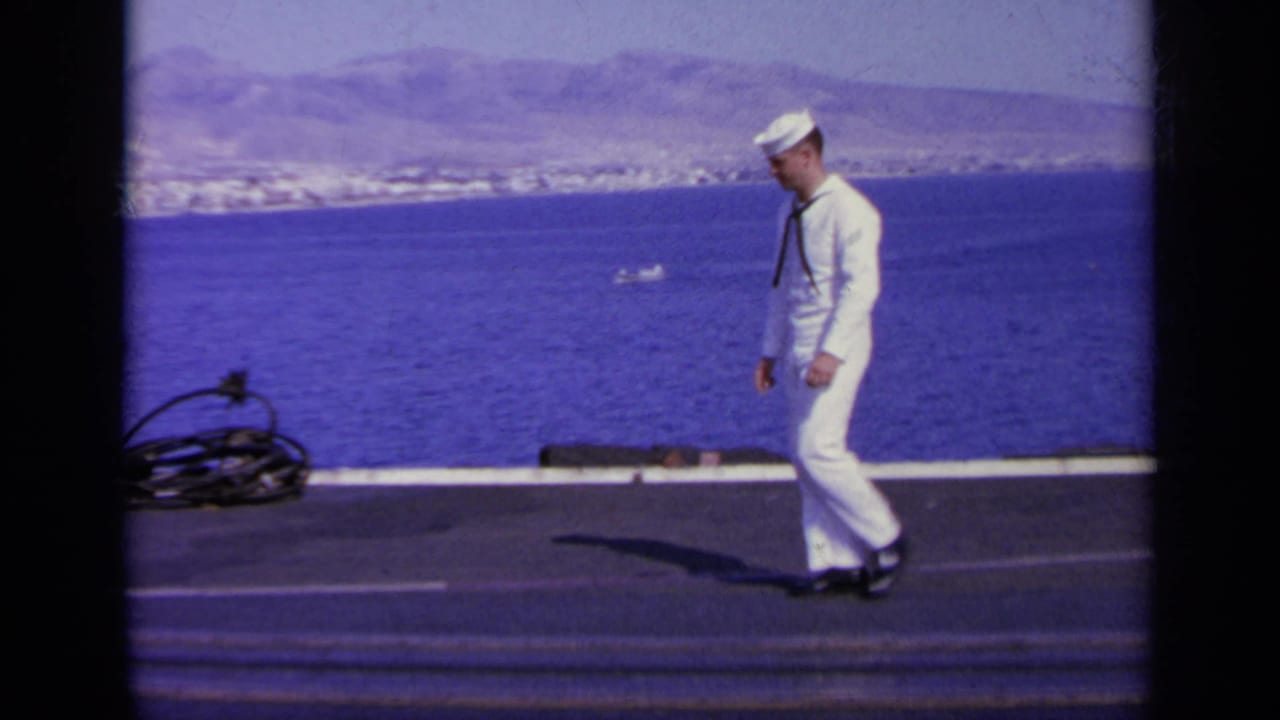Sailor walking across a ship deck beside the sea with a coastal town and mountains in the distance.