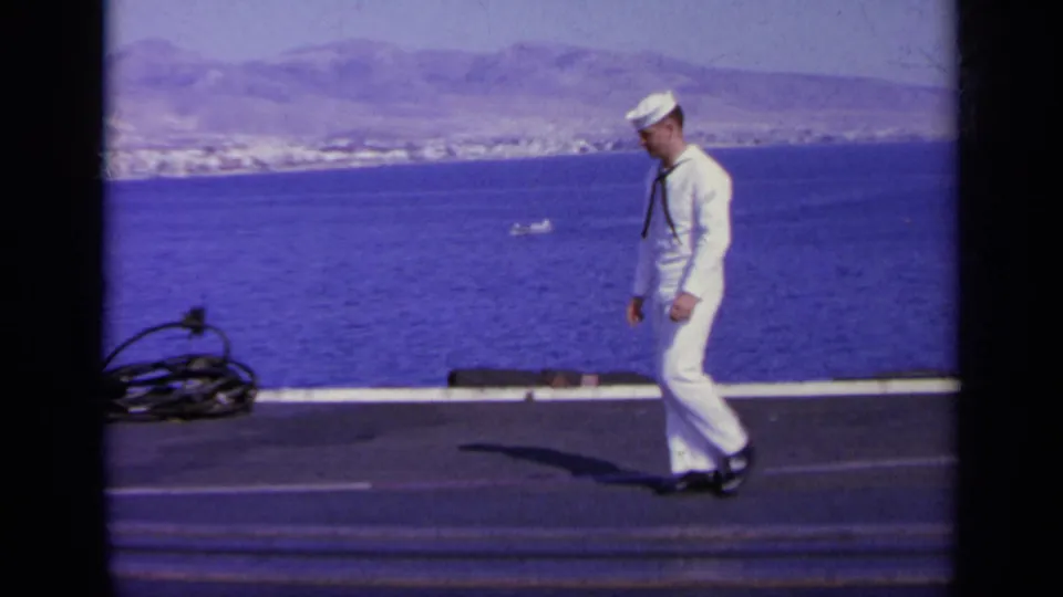 1963 Greece 8mm frame of a sailor in white uniform walking along a ship deck with sea and coastline behind him.