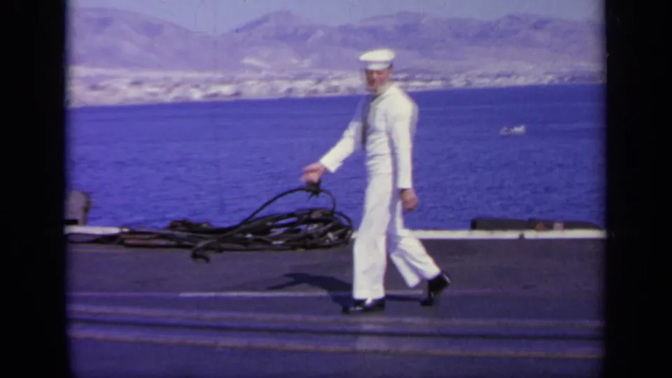 1963 Greece 8mm frame showing a sailor crossing the deck near coiled lines against a blue sea and mountainous coast.