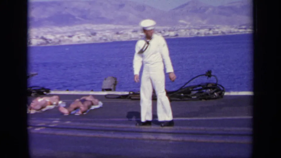 1963 Greece 8mm frame showing a sailor on deck near two sunbathing women, with sea, coastline, and mountains behind them.
