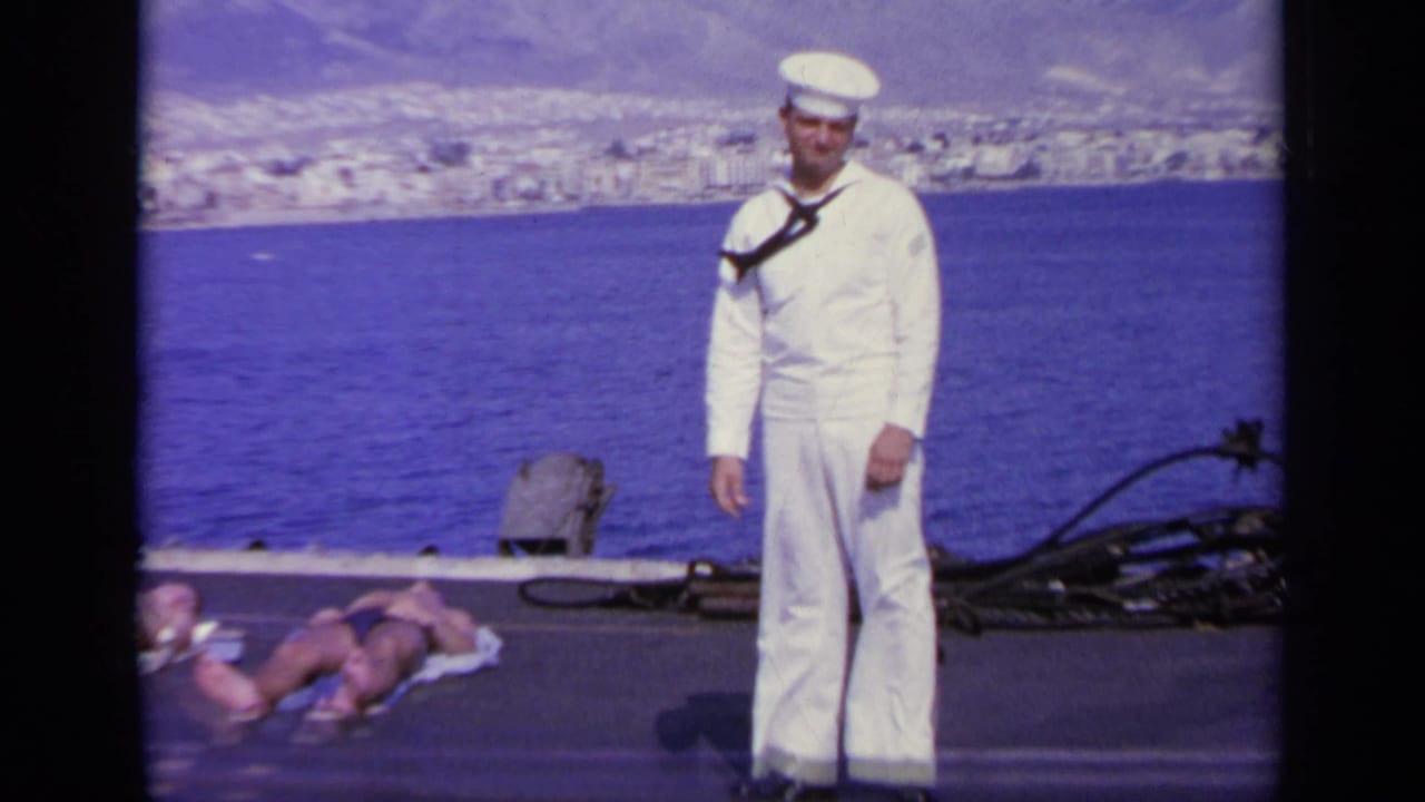 In 1963 Greece, a sailor in white uniform stands on deck before blue water, a coastal town, mountains, and two reclining people.