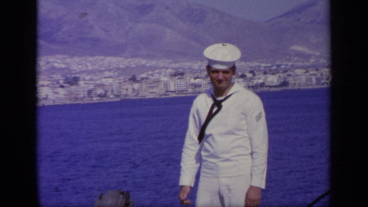 Smiling sailor in white uniform stands near the rail with blue water, town buildings, and mountains behind him.