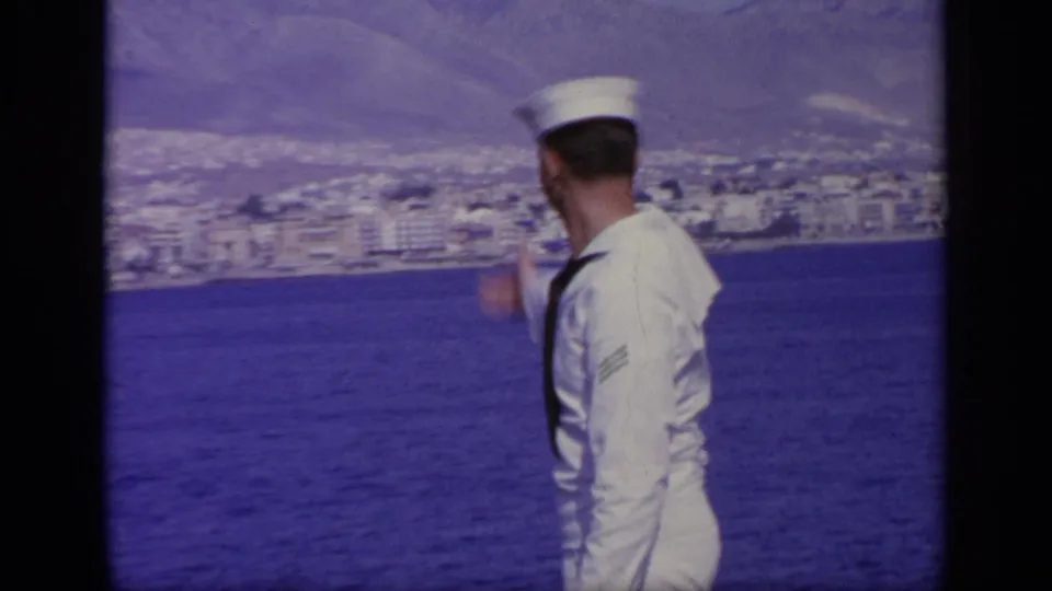 The sailor turns away from the camera and points toward the Greek shoreline, with town buildings and blue water in view.