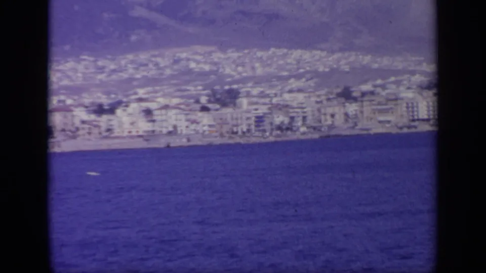 A Greek coastal town and mountain backdrop fill the frame across deep blue water, seen from the ship deck area in 1963 footage.