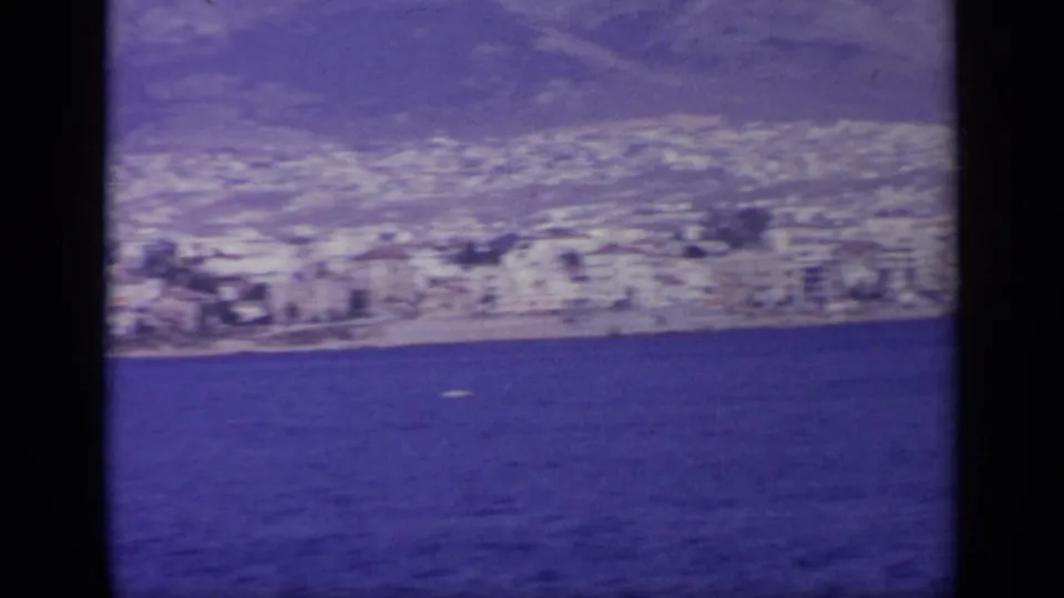 1963 Greece home-movie view of a coastal town and hillside seen from across deep blue water.
