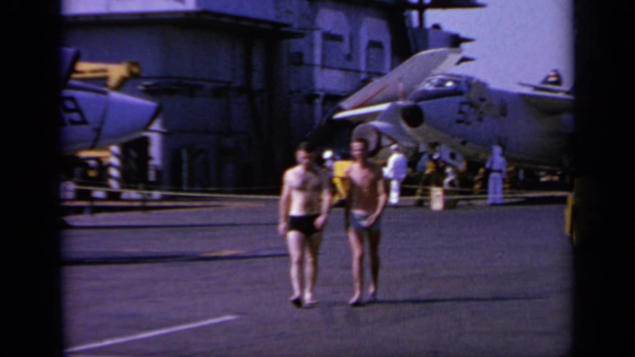 Two men in swim trunks walk on a military aircraft carrier deck in Greece, with parked jets and crew behind them.