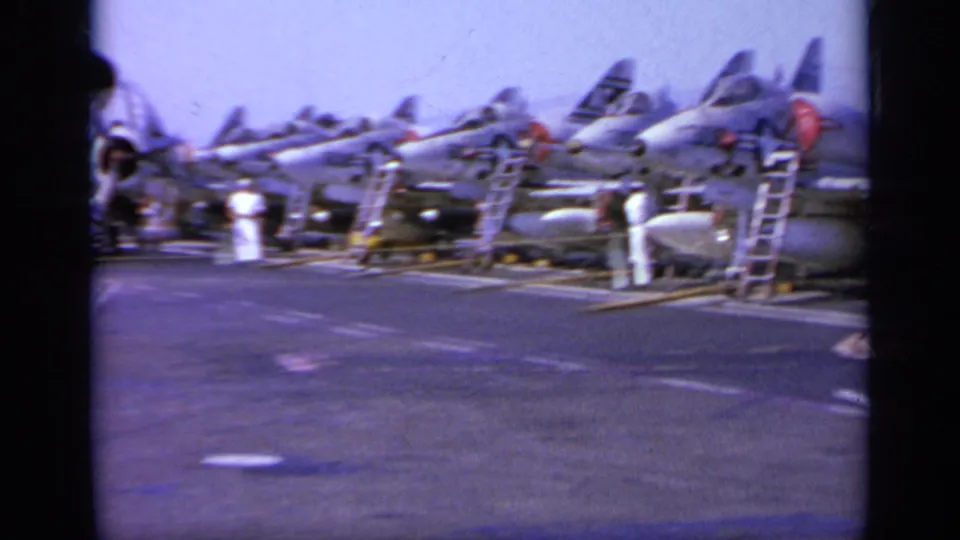 Parked military jets line the aircraft carrier deck while crew in white uniforms work around ladders and equipment.