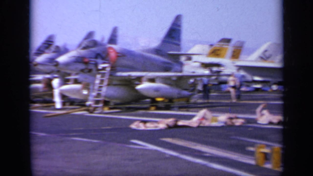 1963 Greece home-movie frame showing men sunbathing on an aircraft carrier flight deck with parked military jets in the background.