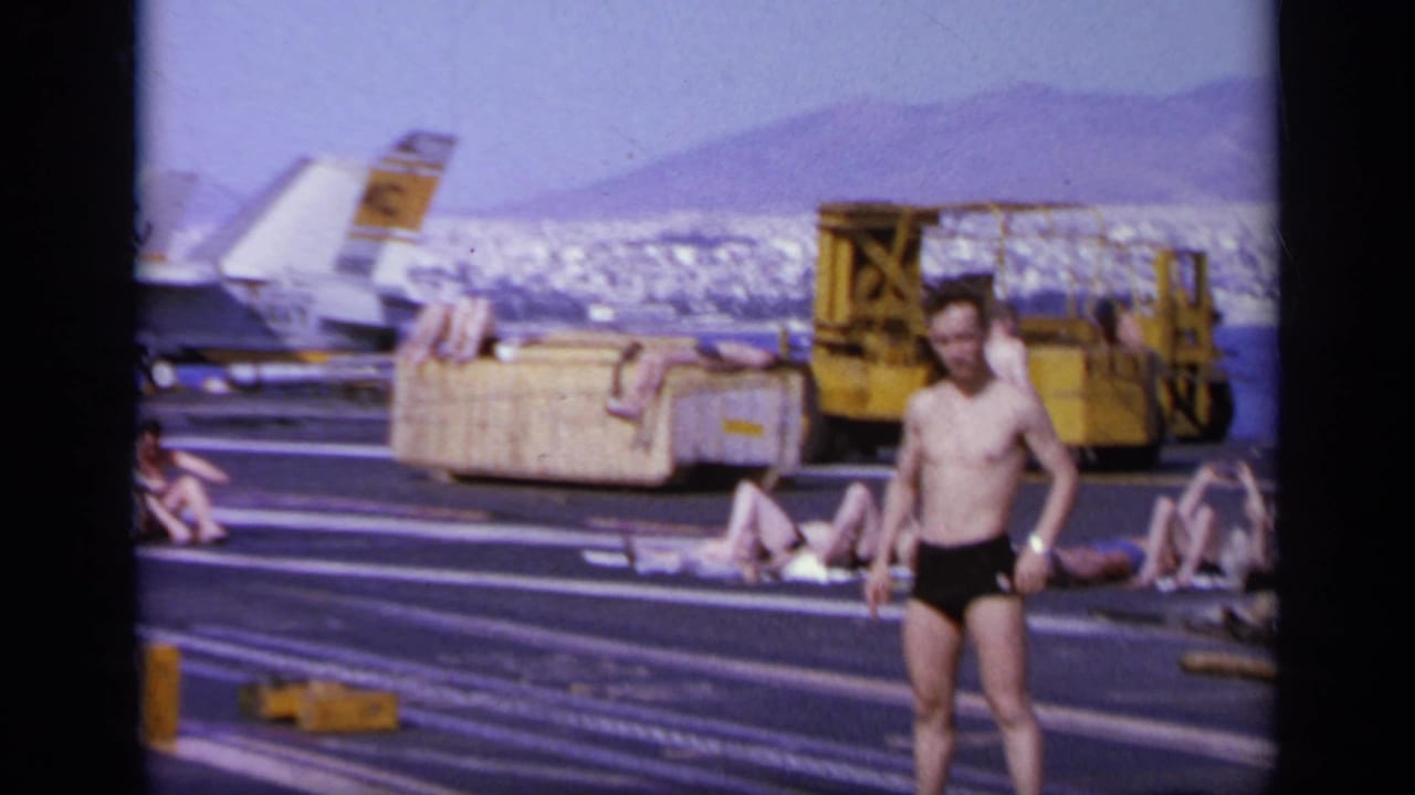 A shirtless man in black trunks waves on the carrier deck near a yellow vehicle.
