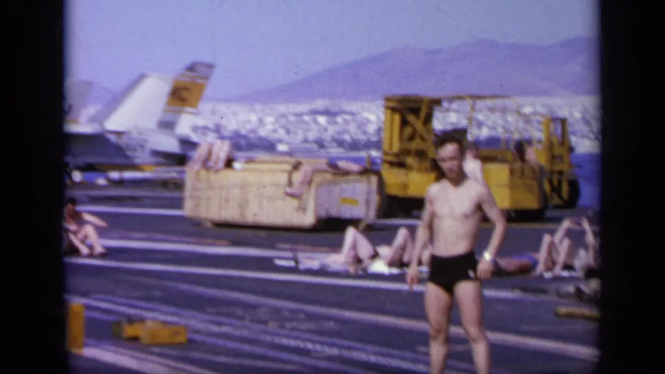 1963 Greece frame of a shirtless man waving at the camera on an aircraft carrier deck, with sunbathers, sea, and deck equipment behind him.