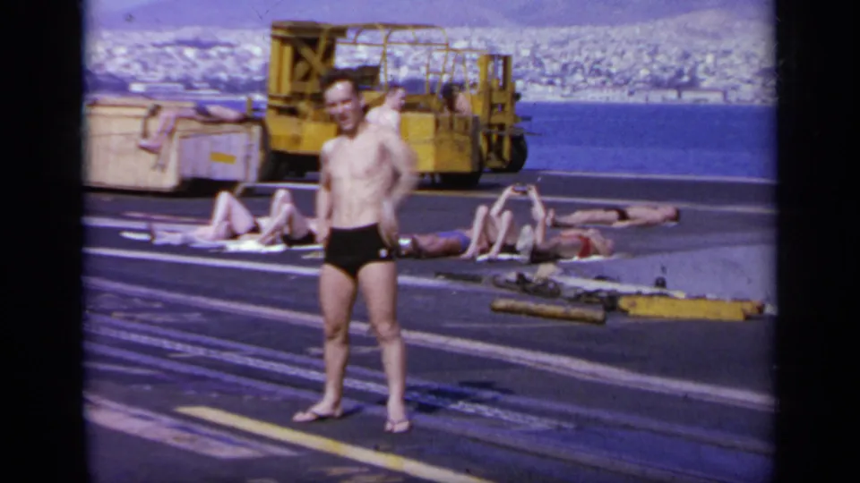 1963 Greece home-movie frame with a man standing in swim trunks on a carrier deck as others lounge near yellow deck equipment and the shoreline.