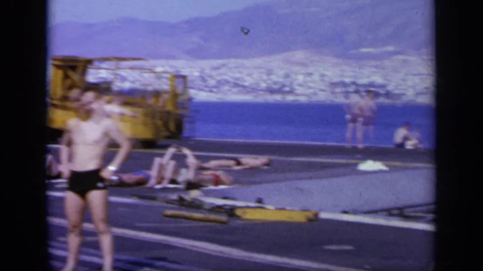 1963 Greece frame showing men sunbathing on an aircraft carrier deck, with yellow equipment, blue water, mountains, and a coastal city in view.