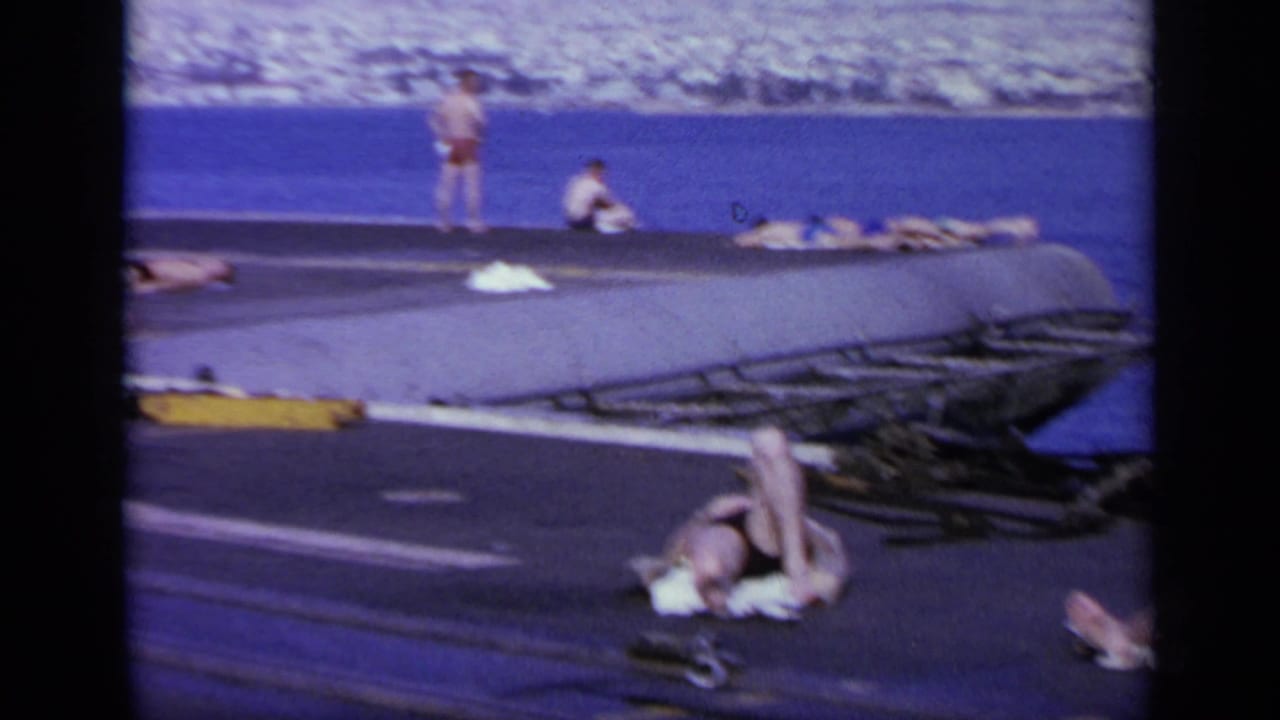 1963 Greece home-movie frame showing people in swimwear resting on a ship deck with sea and coastline beyond.