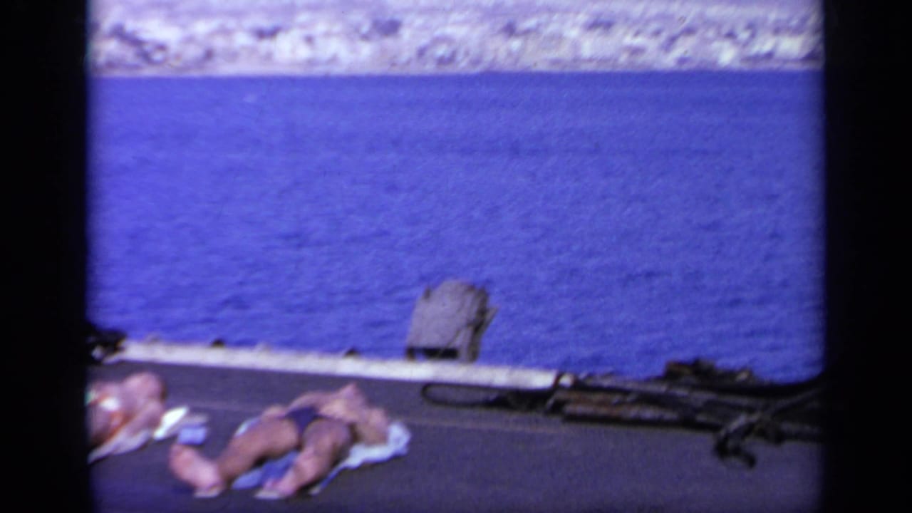 Passengers sunbathe on the deck of a vessel in Greece, with blue sea and a pale distant shore in the background.