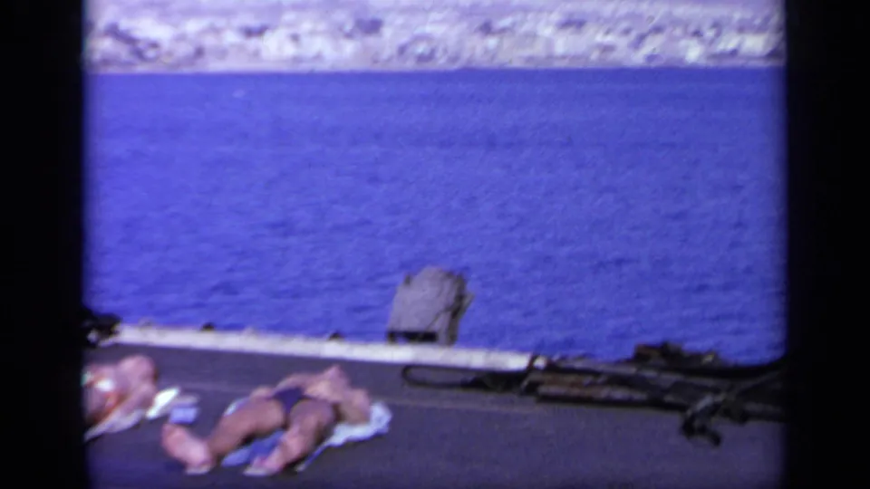 Passengers sunbathe on the deck of a vessel in Greece, with blue sea and a pale distant shore in the background.