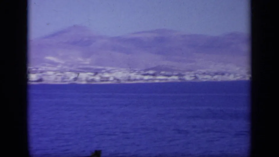 1963 Greece 8mm frame looking from the ship across the sea toward a mountainous coastal settlement.