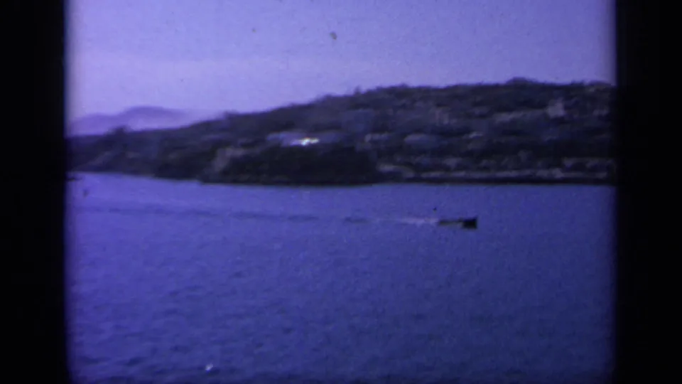 A shipboard view in Greece shows blue water, a small boat, and a dark mountainous coast under a hazy sky.