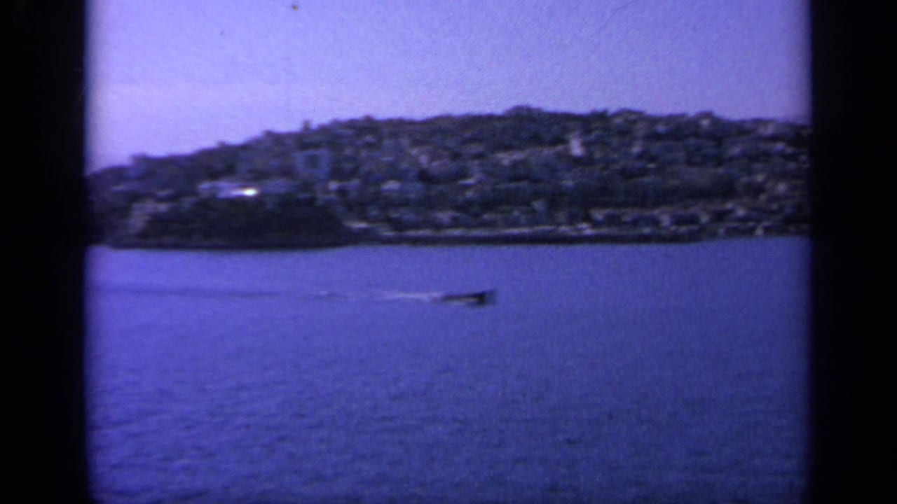 Coastal hillside town across blue water with a small boat wake in the foreground.