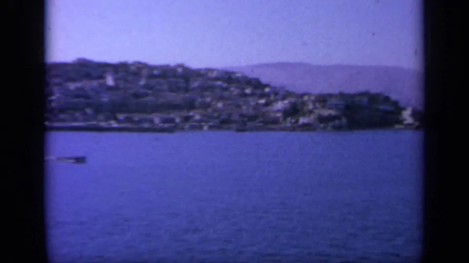 1963 Greece 8mm frame with a built hillside coast, open blue water, and dark vertical film-frame borders.