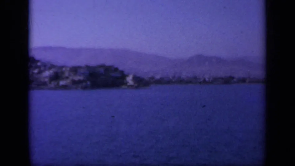 1963 Greece 8mm frame looking across blue water toward a low shoreline and hazy mountain forms.