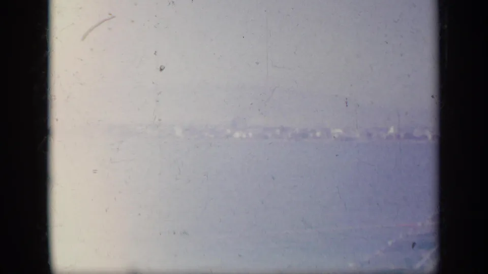 Soft 8mm frame of a Greek coastal view, with faint buildings along the shore and mountains beyond the water.