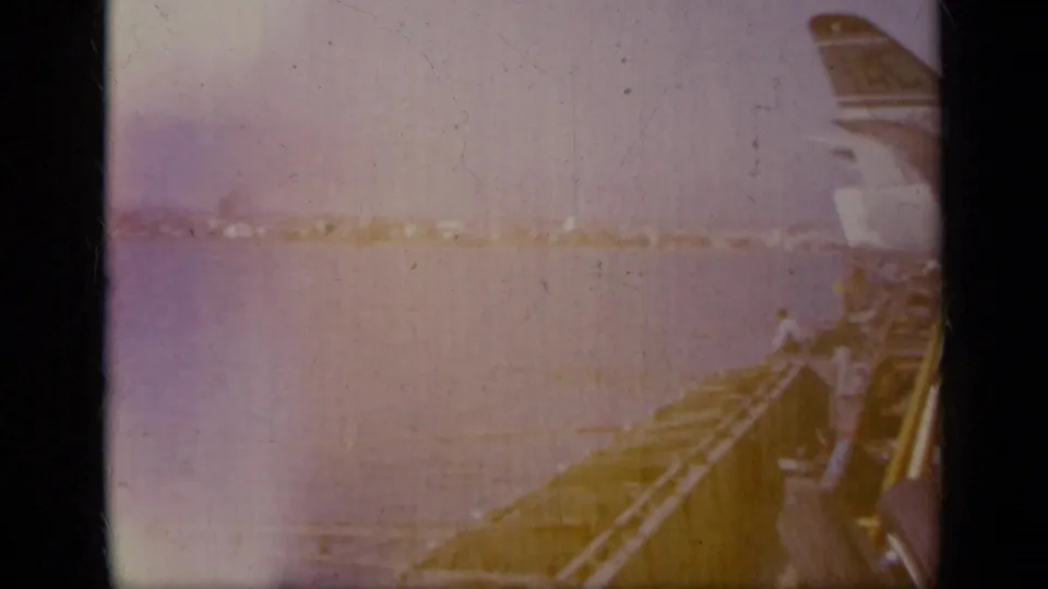 1963 Greece home-movie frame looking past a pier or vessel edge toward water, shoreline and hazy mountains.