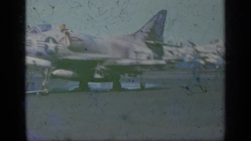 Faded 1963 8mm frame from Greece showing a U.S. Navy jet parked on an aircraft carrier deck with personnel and equipment nearby.