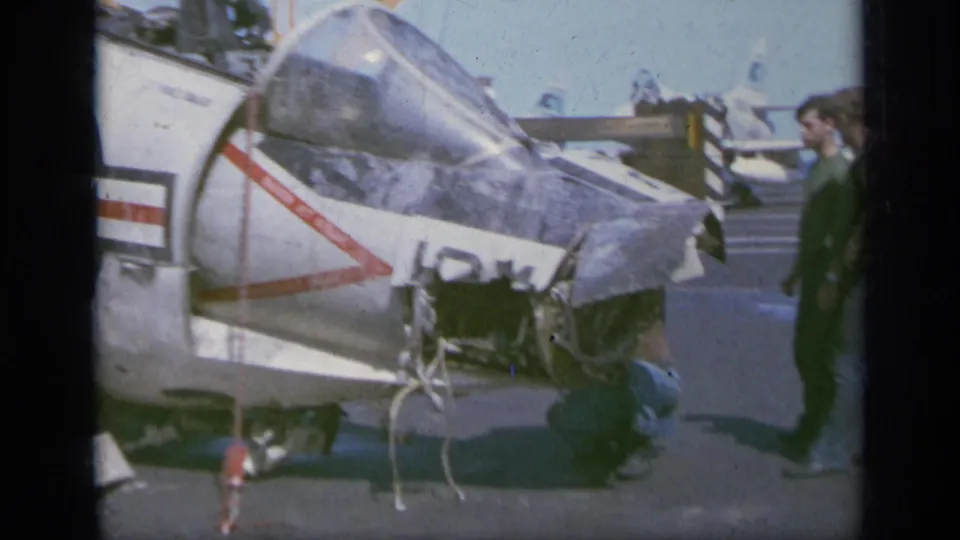 1963 Greece 8mm frame showing a parked military-style aircraft with cockpit canopy, painted tail markings, landing gear, and nearby people.