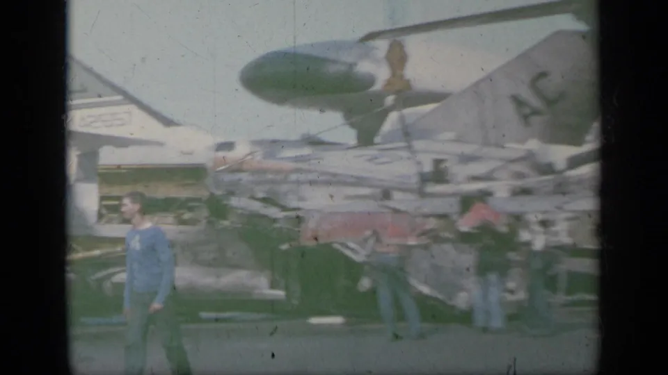 1963 Greece 8mm frame showing crew or visitors near white U.S. jet fighters on a carrier deck, with bright sky and aircraft noses visible.