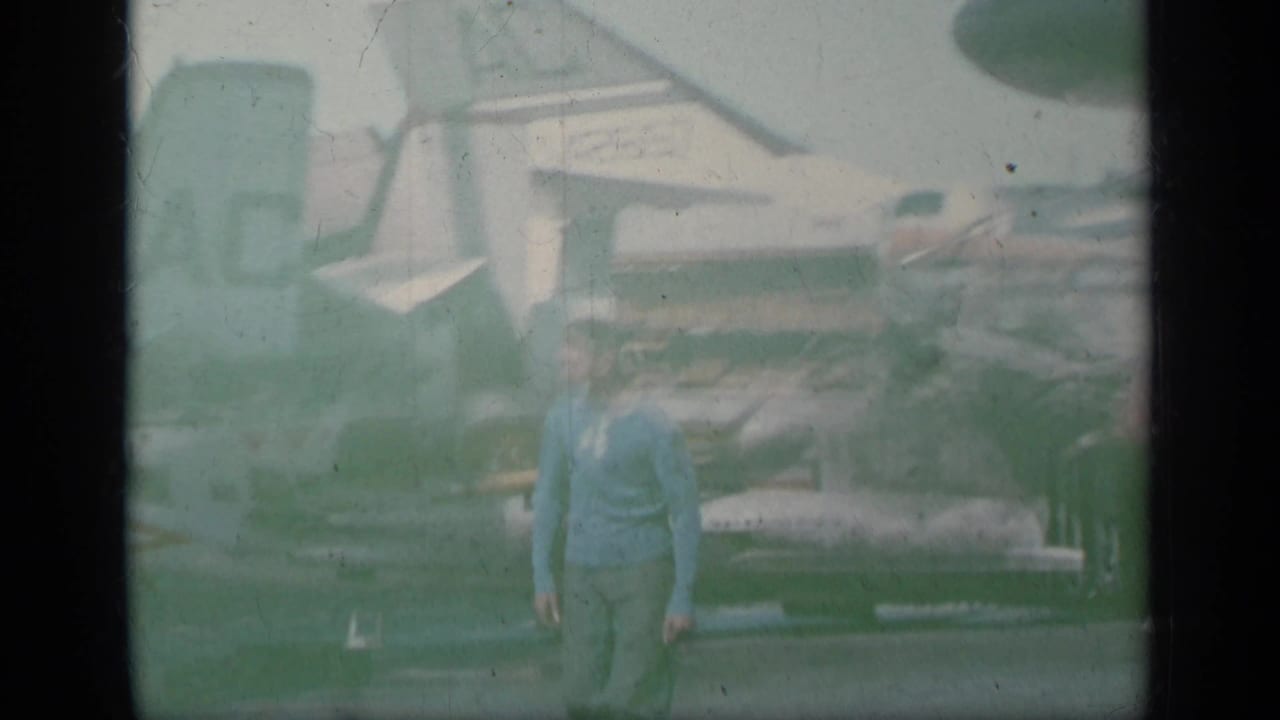 Man in a blue shirt walking on a carrier deck near parked jets.