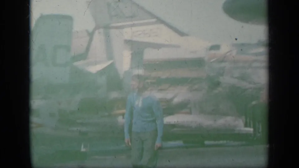 1963 Greece 8mm frame showing a man in a blue shirt on an aircraft carrier deck with parked U.S. Navy jets behind him.