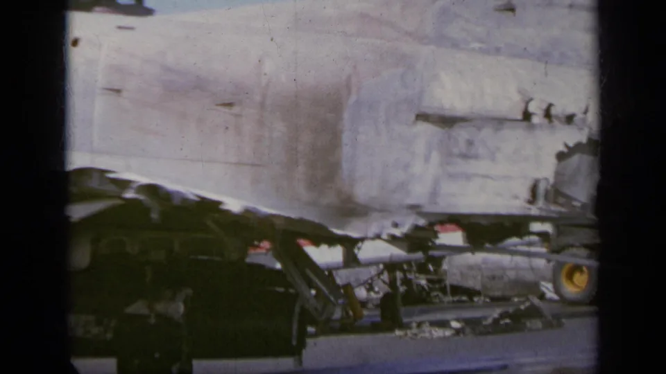 1963 Greece 8mm frame showing the damaged side of a jet on a carrier deck with equipment and small crew figures nearby.