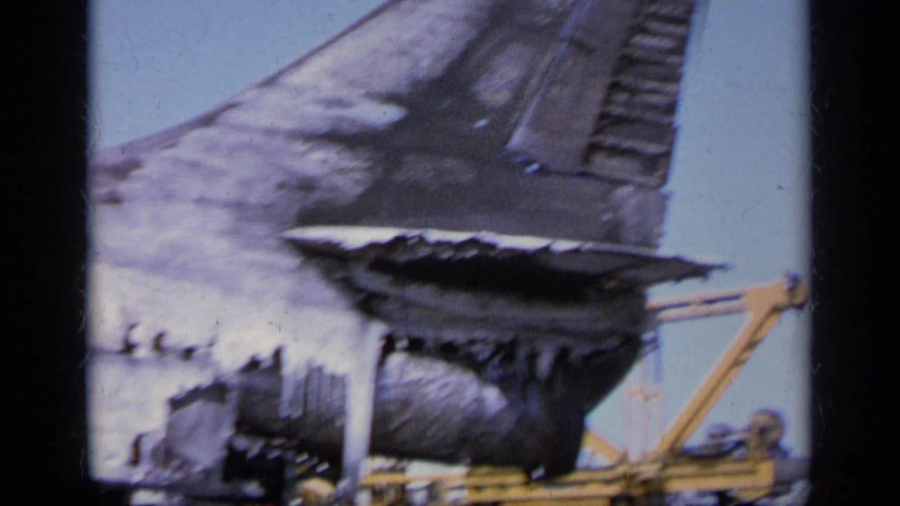 Burned aircraft tail section with yellow ground equipment on a bright deck.